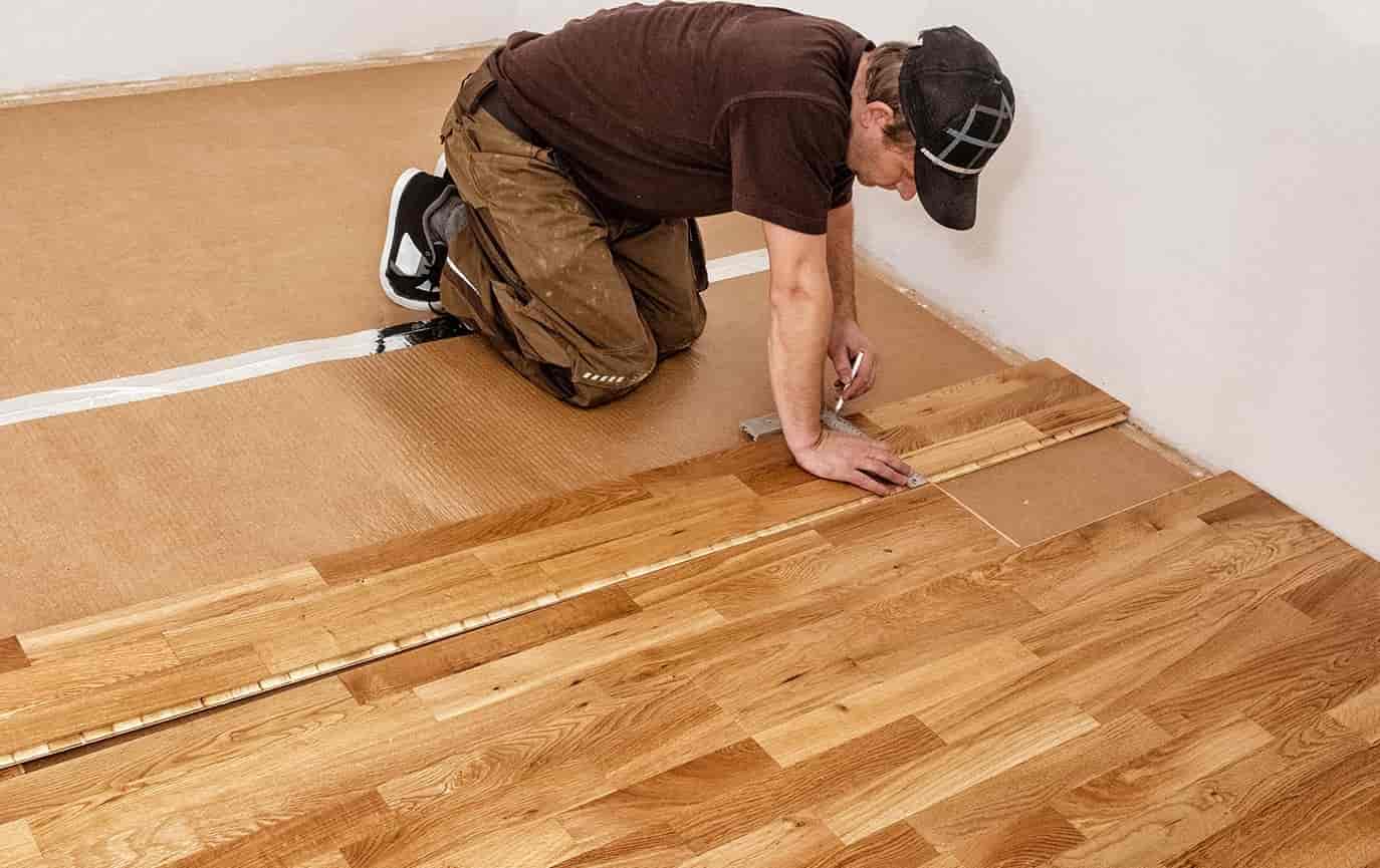 picture showcasing the installation of engineered wood flooring. A man in work wear is on his hands and knees in a half floored room, installing the flooring.